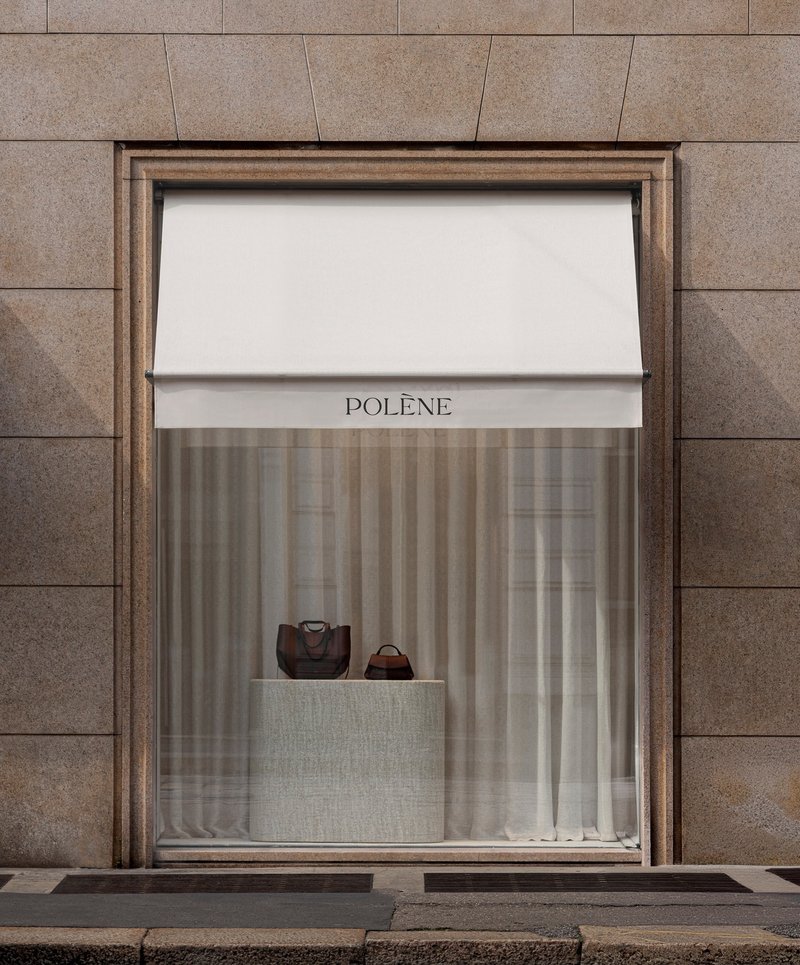 Single window display framed in bronze with white awning and sheer curtains against stone panels