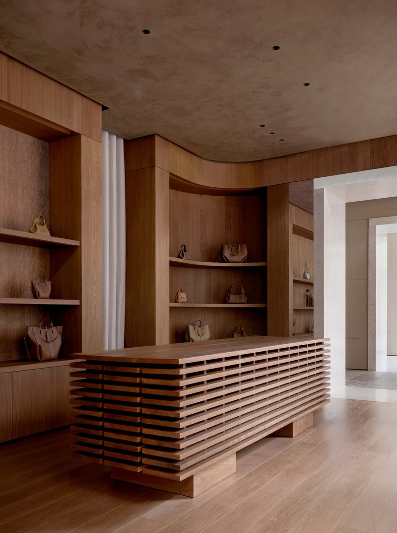 Timber display counter with horizontal slat base and curved shelving wall under a limestone ceiling