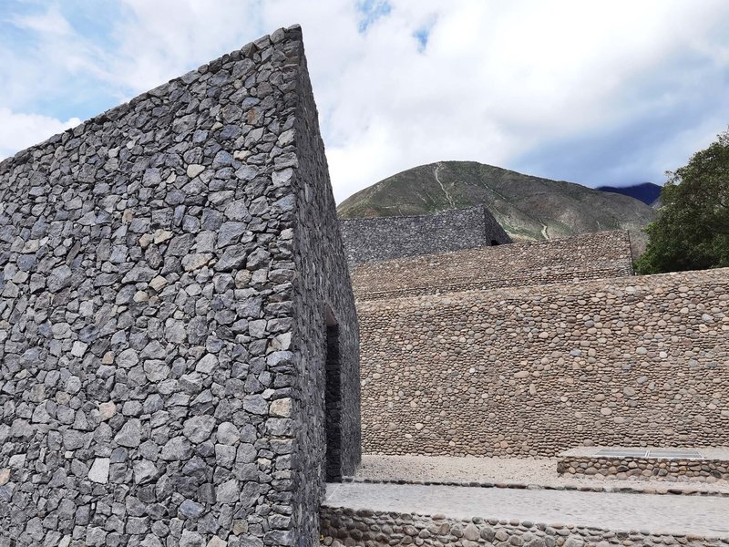 Angled stone wall made of irregular grey rocks set against a hillside under partly cloudy sky