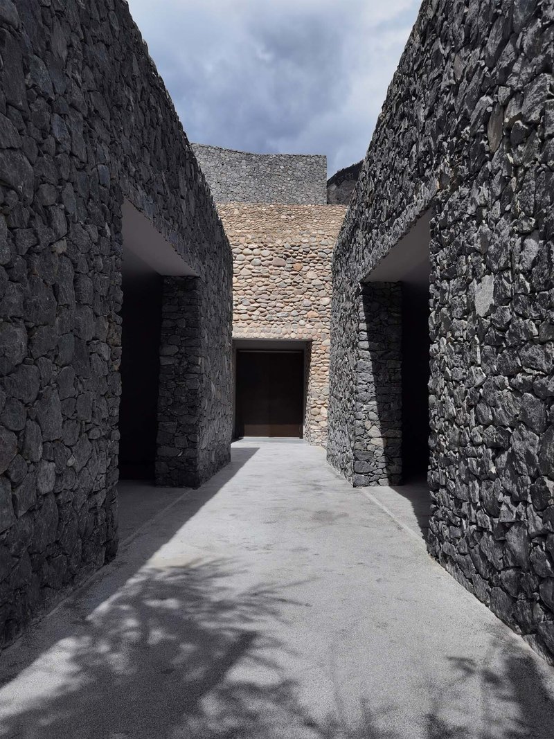Narrow outdoor passageway between tall stone walls leading to a central opening with dappled shadows