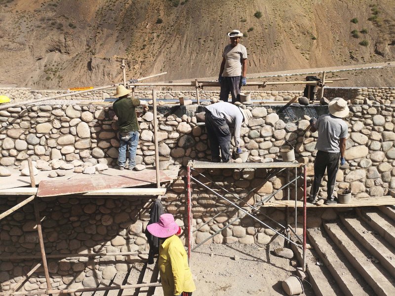 Construction workers on scaffolding assembling a river stone retaining wall beneath a mountain