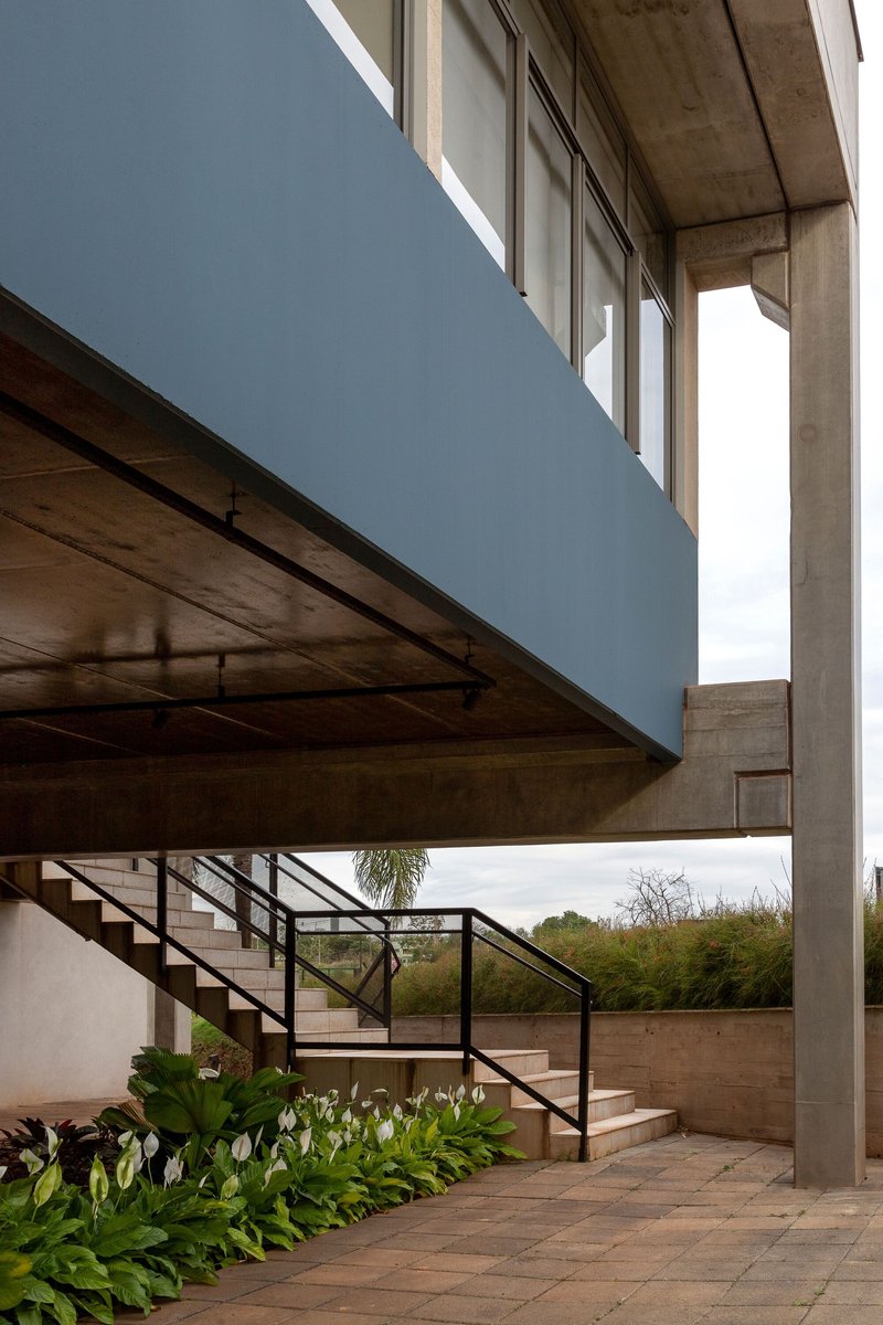 Exterior stair with metal handrails ascending beneath a cantilevered blue volume and exposed concrete structure