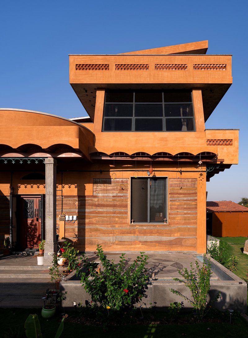 Terracotta brick facade with cantilevered upper volume, scalloped overhangs, and potted plants in afternoon light