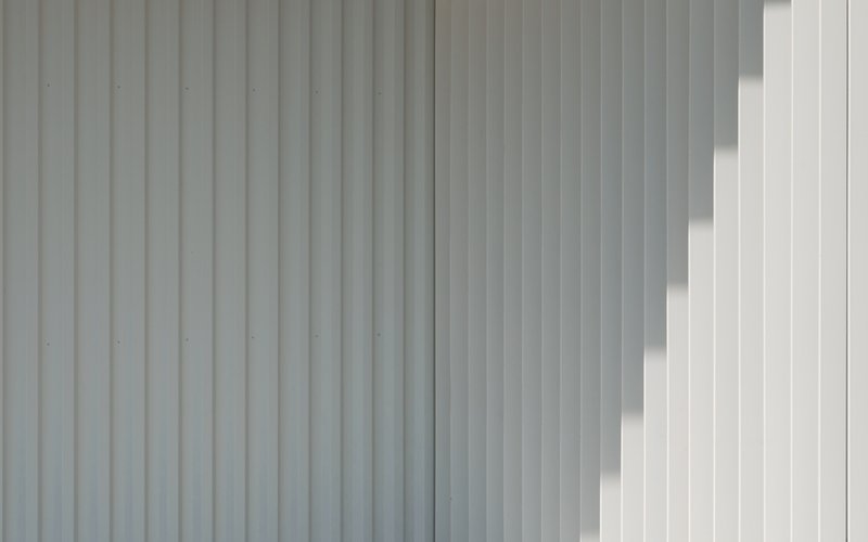 Close-up of vertical corrugated metal cladding in light grey with diagonal sunlight shadows across the surface