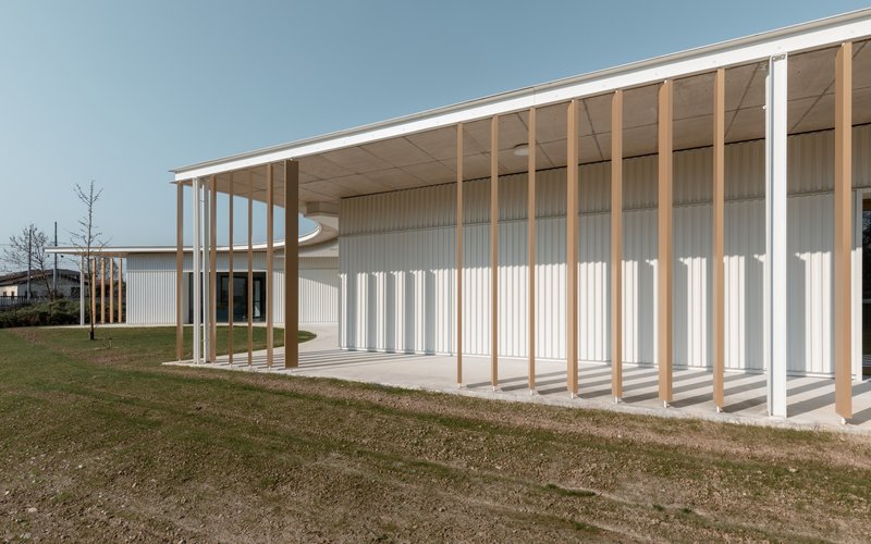 Covered walkway with timber columns and exposed plywood ceiling framing the corrugated facade and lawn in late afternoon