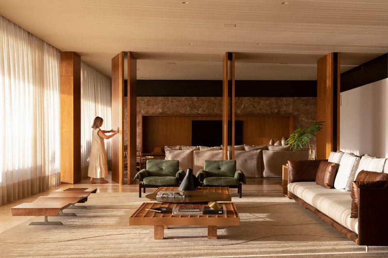 Living room with timber columns and slatted ceiling as a figure adjusts sheer curtains near the glazed wall