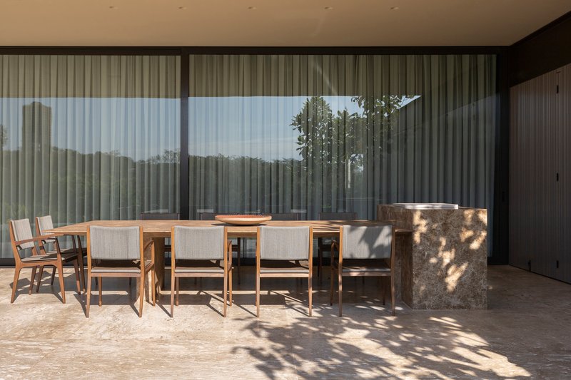 Covered terrace with dining table and chairs casting dappled shadows under tree canopy beyond glass