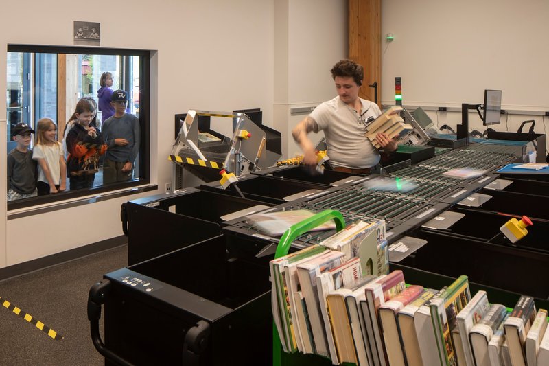 Automated book sorting conveyor belt with green bins and onlookers visible through a glass window