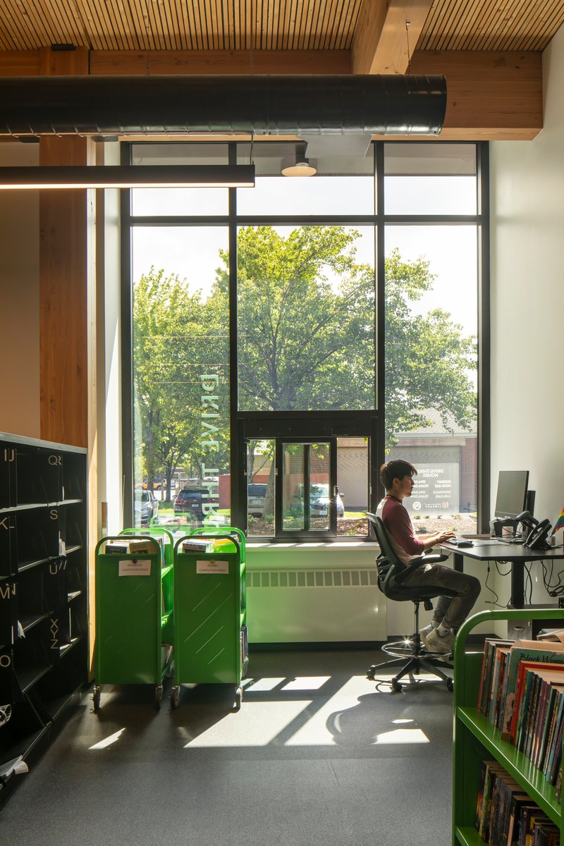 Workstation beneath a steel beam and timber ceiling with green bins and tall windows overlooking street trees