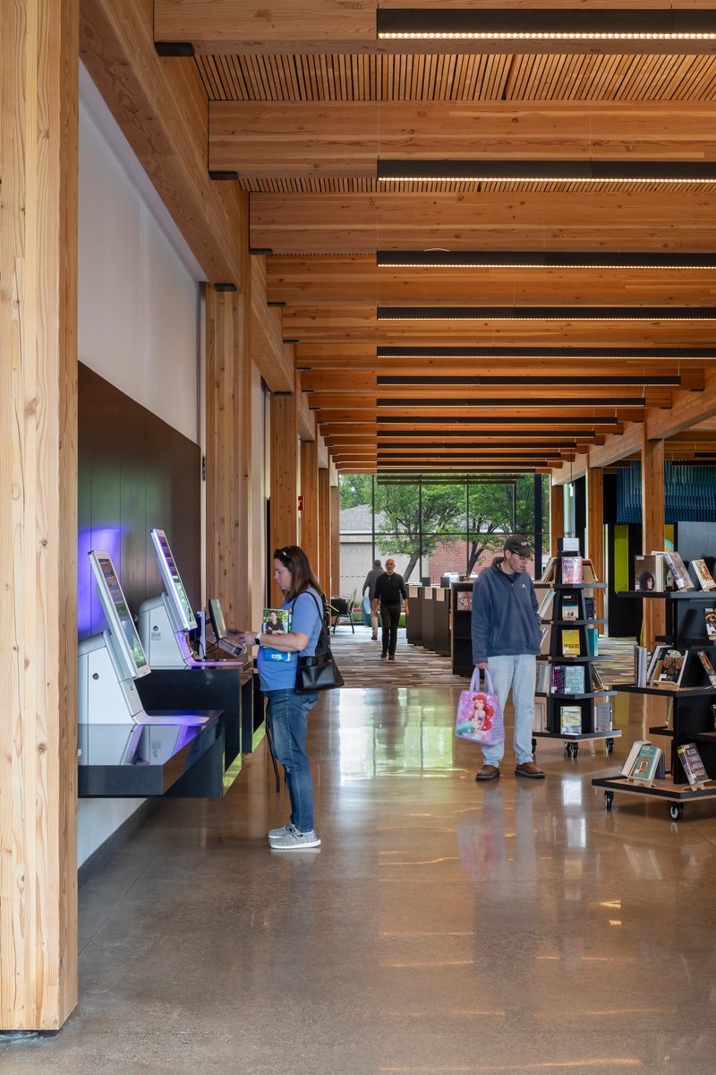 Long library corridor with rhythmic timber beams and ceiling slats as patrons use digital catalog stations