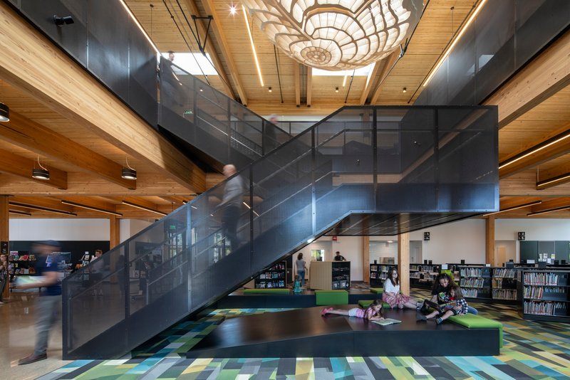Double-height library interior with dark metal staircase, timber ceiling beams and children seated on multicolored floor cushions