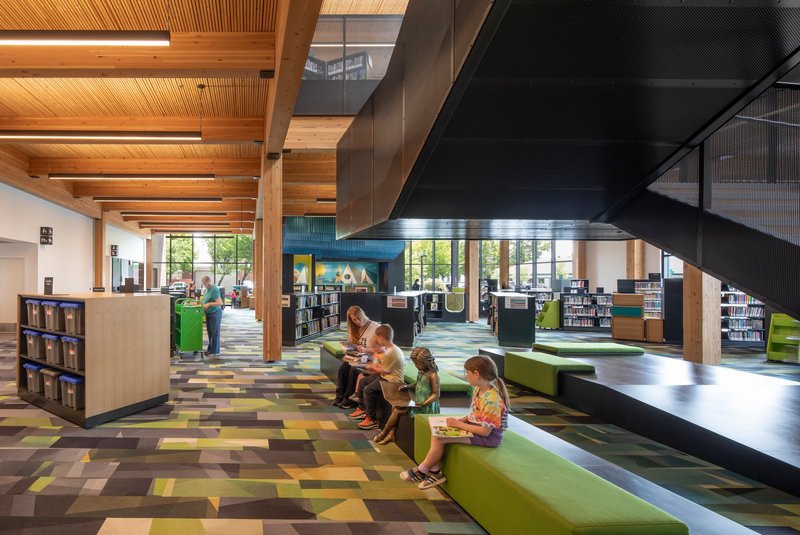 Library reading area with green stepped seating platform beneath a suspended metal staircase and exposed timber ceiling