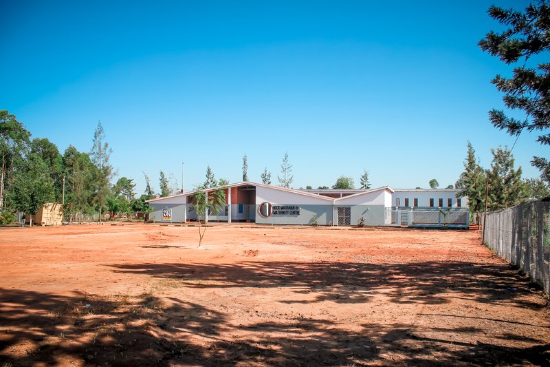 Wide view of the single-story complex across a cleared red dirt courtyard with young trees