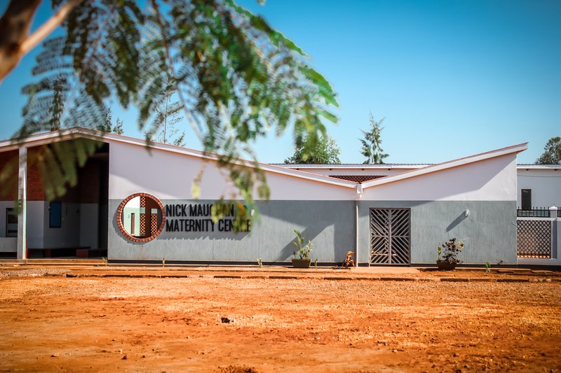 Front facade with butterfly roof and circular logo seen through overhanging foliage in midday sun