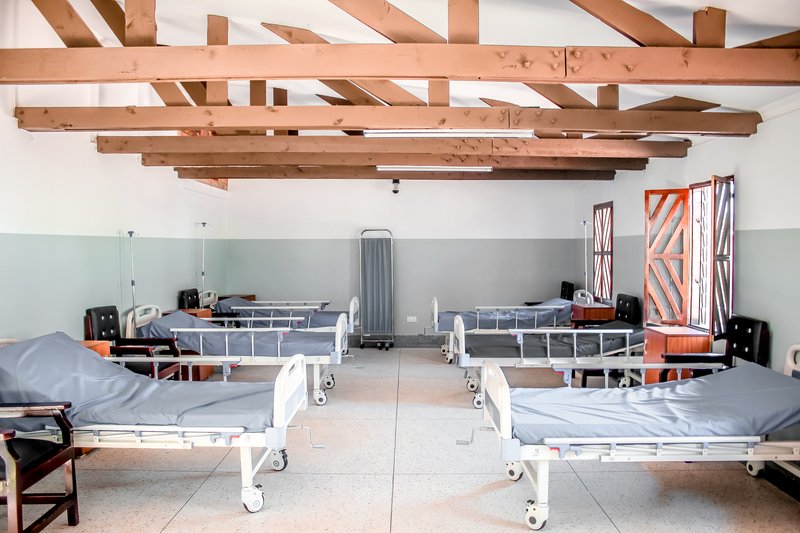 Interior ward with metal hospital beds beneath an exposed timber beam ceiling and grey wainscoting