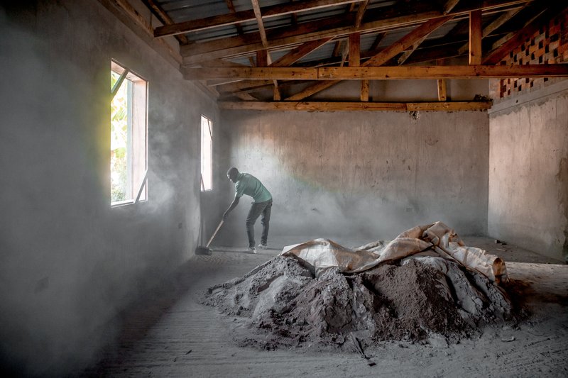 Interior room under construction with a worker shoveling cement amid dusty air and exposed timber roof trusses