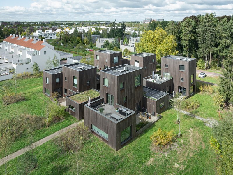 Aerial view of dark timber-clad volumes with roof terraces arranged across a green lawn with scattered young trees