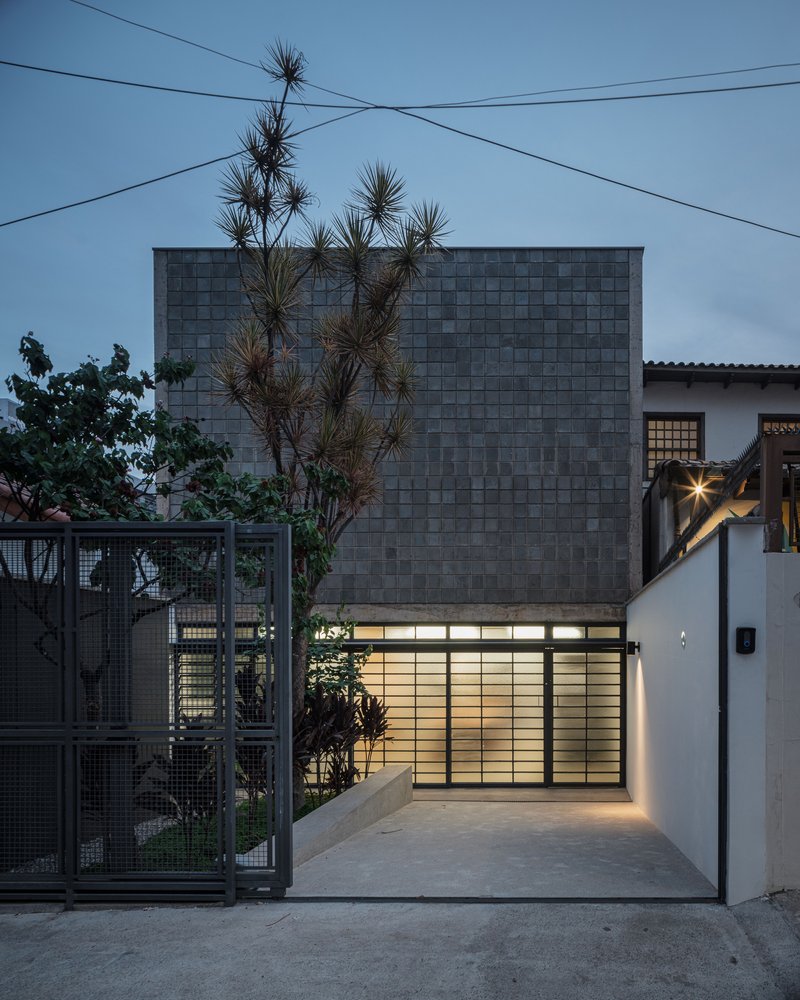 Street facade with dark ceramic tile upper volume, backlit glass block entry, and spiky yucca tree at dusk