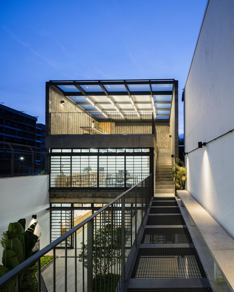 Exterior metal staircase ascending past translucent glazed walls and a glazed roof canopy at dusk