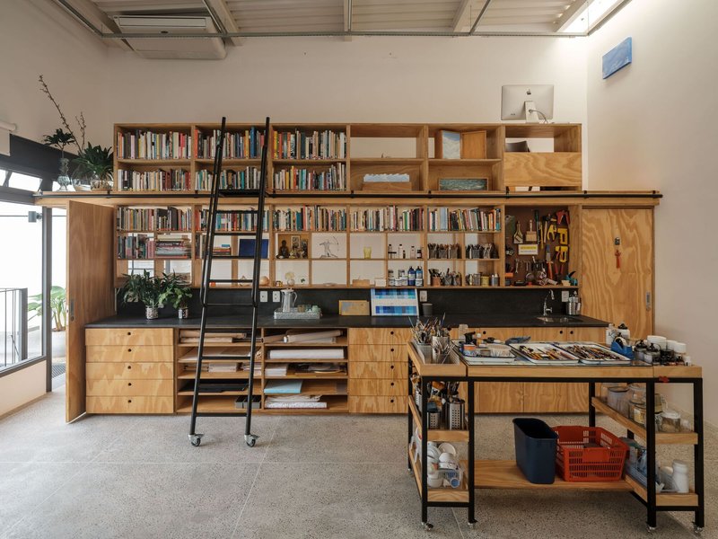 Floor-to-ceiling plywood storage wall with rolling ladder and black steel work table in foreground