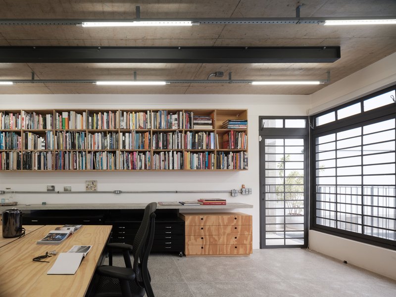 Wall-mounted timber bookshelves above built-in desk with gridded window screen and exposed concrete beams