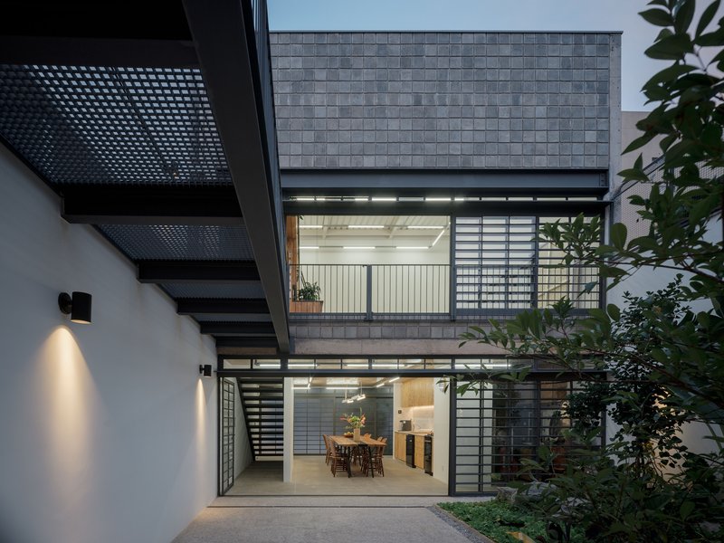 Courtyard facade at dusk with gray tile cladding, gridded screens, perforated metal stairs, and planted garden