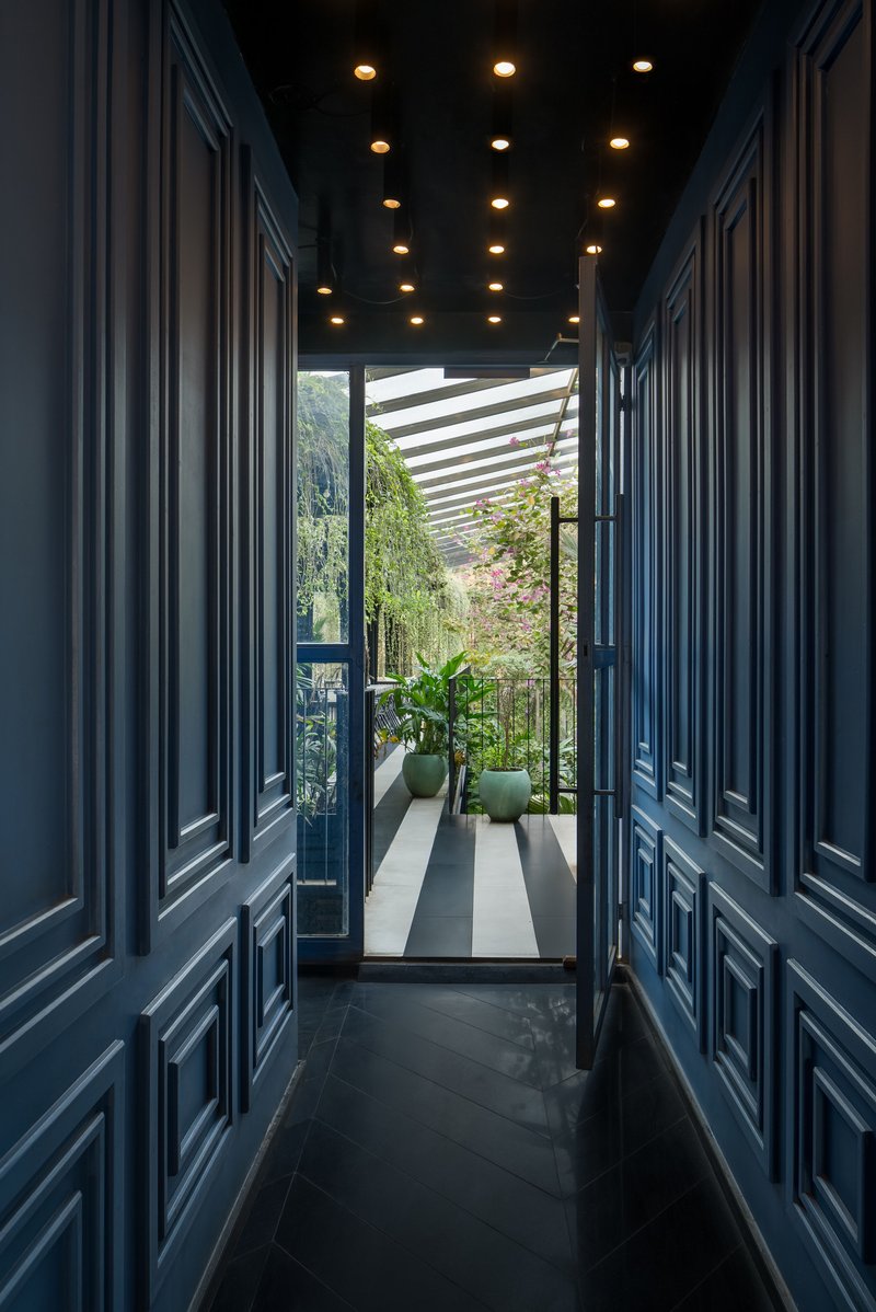 Narrow corridor with navy paneled walls and ceiling-mounted downlights leading to a planted terrace with slatted pergola