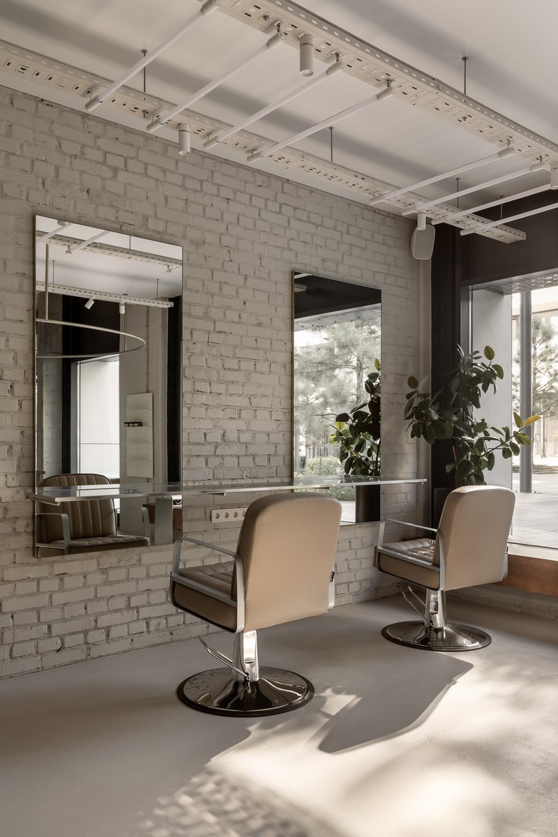 Salon area with white-painted brick wall, framed mirrors, styling chairs, and sunlight filtering through glass