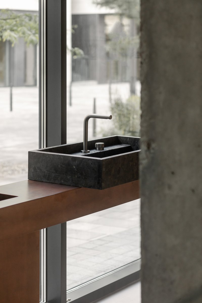 Close-up of dark stone basin with matte black faucet on timber shelf beside concrete column