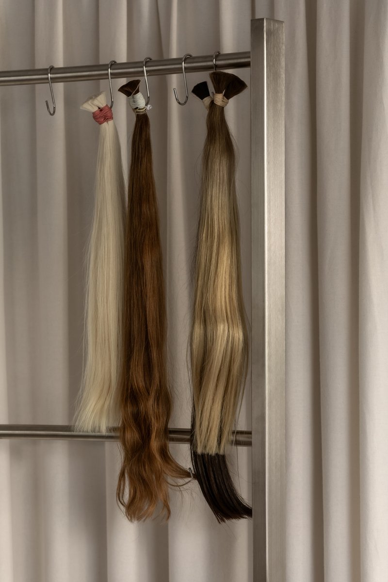Three bundles of hair extensions hanging from a metal rail against a pale curtain backdrop