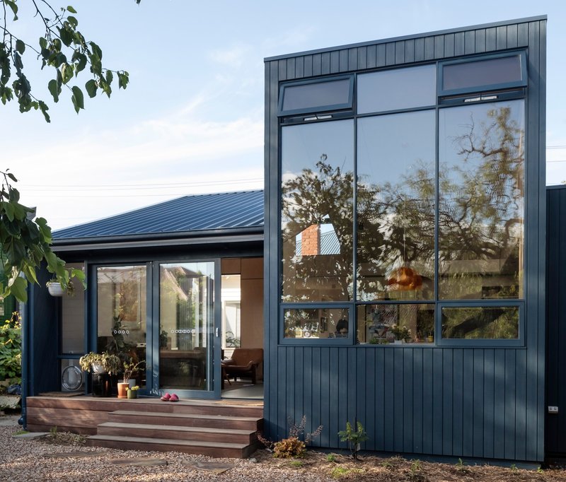 Two-volume residence with vertical metal cladding and tall glazing beside timber entry steps and gravel courtyard