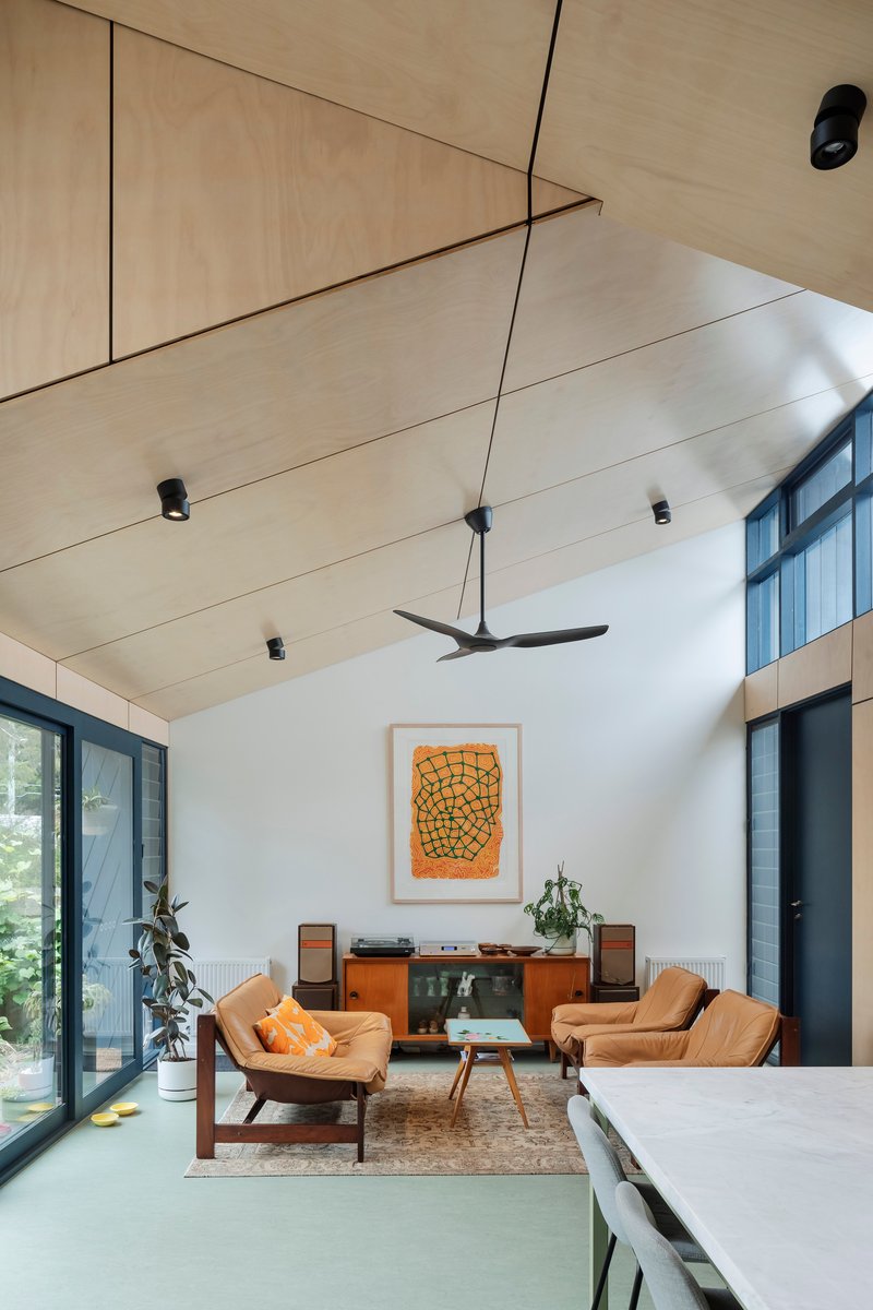 This architectural image showcases a modern living room with a vaulted plywood ceiling and abundant natural light. The space features mid-century modern furnitu