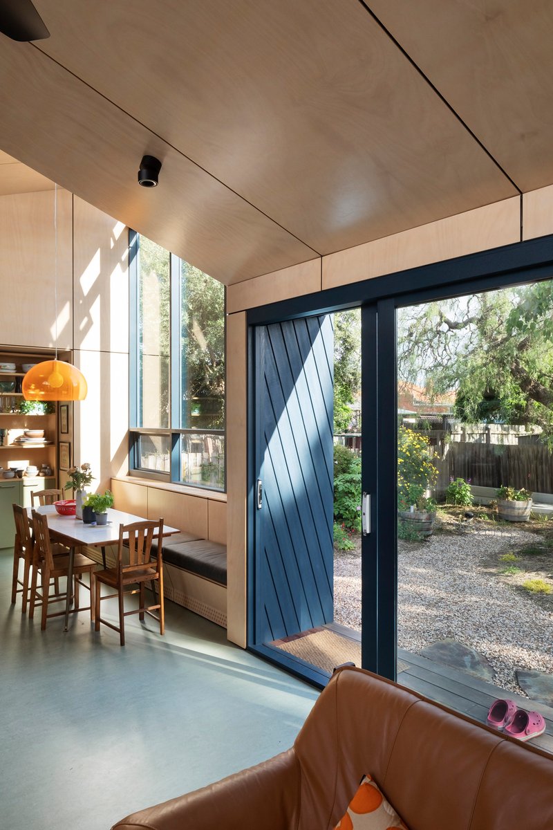 This architectural image showcases a modern interior with a focus on natural light and indoor-outdoor connection. The space features plywood paneling, a dining