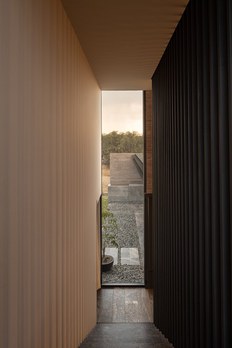 Vertical slit window between timber and black slat walls opening to a gravel courtyard