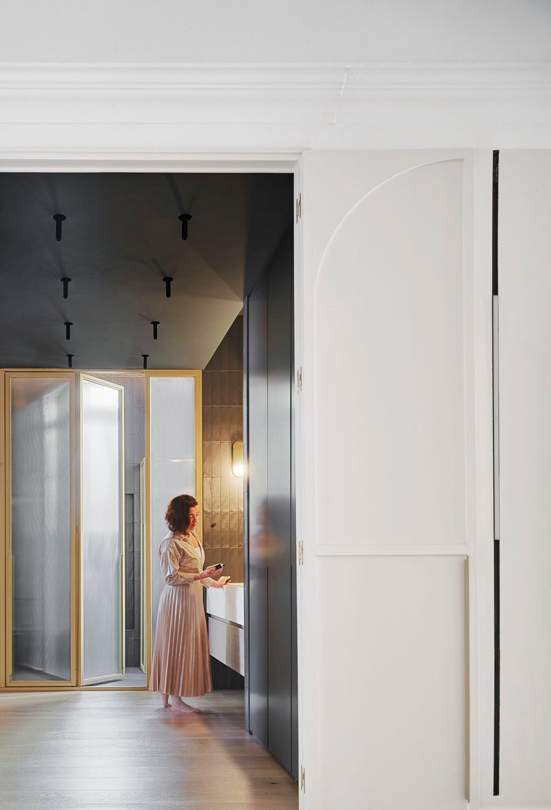 Woman in a pleated dress standing beside dark grey cabinetry and brass-framed translucent glass doors