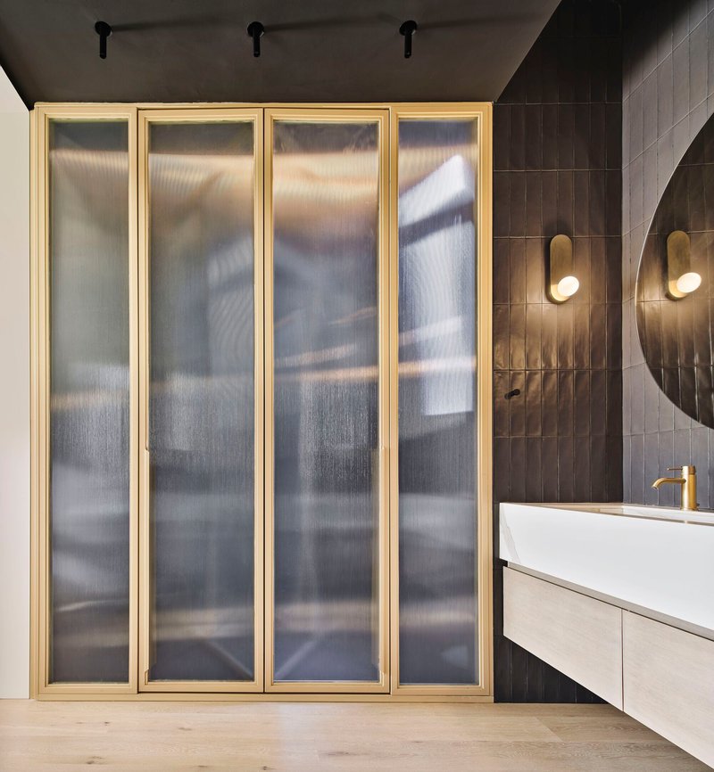 Brass-framed translucent glass folding doors adjacent to dark tiled bathroom wall with circular mirror and sconces