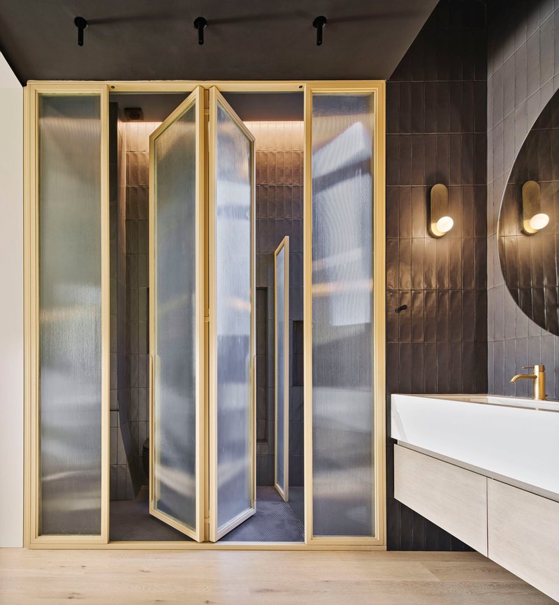 One panel of the brass-framed glass partition opening to reveal the dark tiled shower beyond