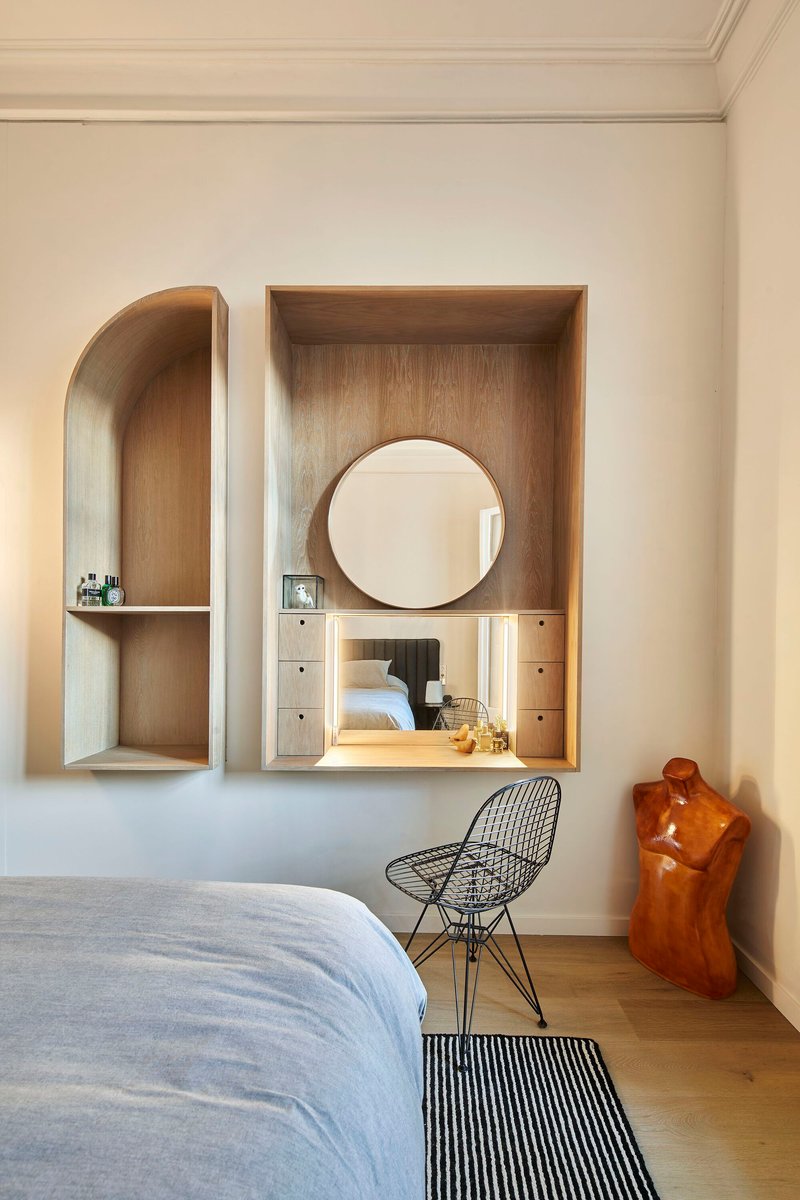 Built-in timber vanity niche with round mirror and arched wall shelf beside wire chair and carved wood sculpture