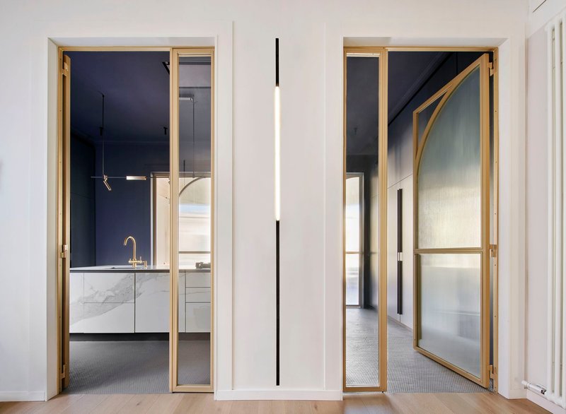 Brass-framed pivoting door with frosted glass panel and arched structural brace leading to kitchen beyond
