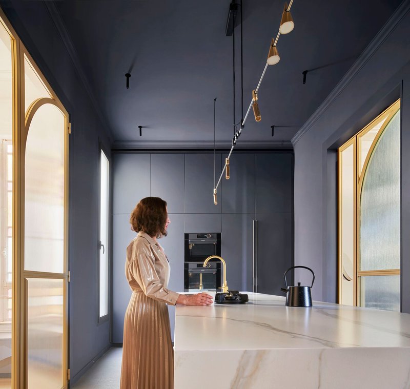 Kitchen with slate blue cabinetry and marble island where a person stands by the brass faucet