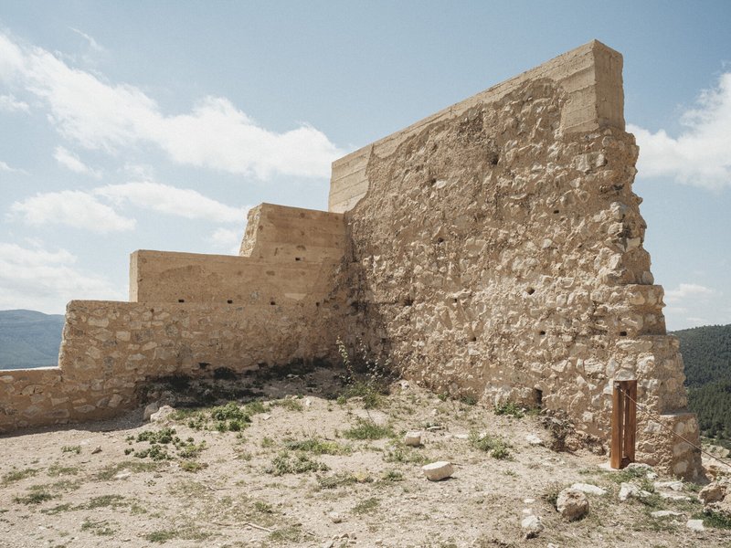 Reconstructed tower corner with layered stone masonry stepping down a bare hillside under partly cloudy skies