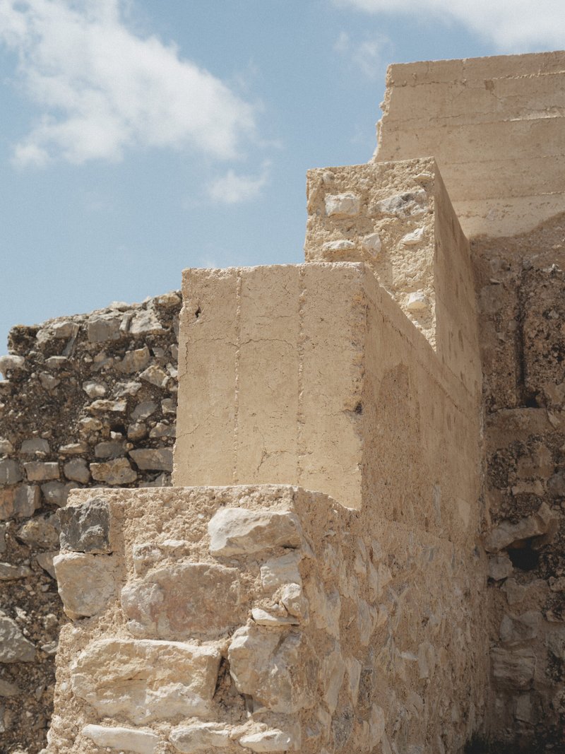 New rammed earth column meeting weathered stone masonry wall under clear sky