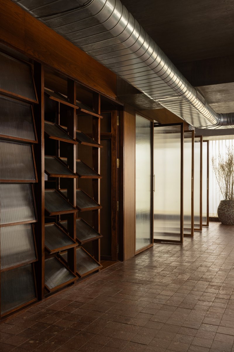 Corridor with exposed ductwork ceiling and geometric wooden shelving beside translucent glass partitions