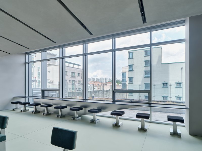 Row of dark upholstered stools facing floor-to-ceiling windows with views of surrounding residential buildings