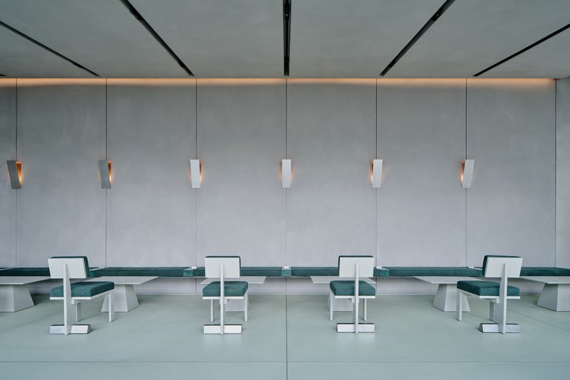 Symmetrical wall of grey panels with six suspended pendant lights above teal upholstered benches and white chairs