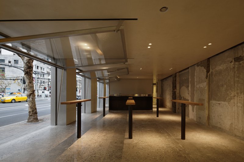 Ground floor interior with street trees visible through glass facade and standing height work tables on terrazzo flooring