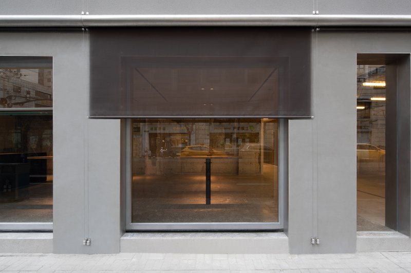 Street facade with gray metal panels and retractable mesh screens above the glazed storefront opening