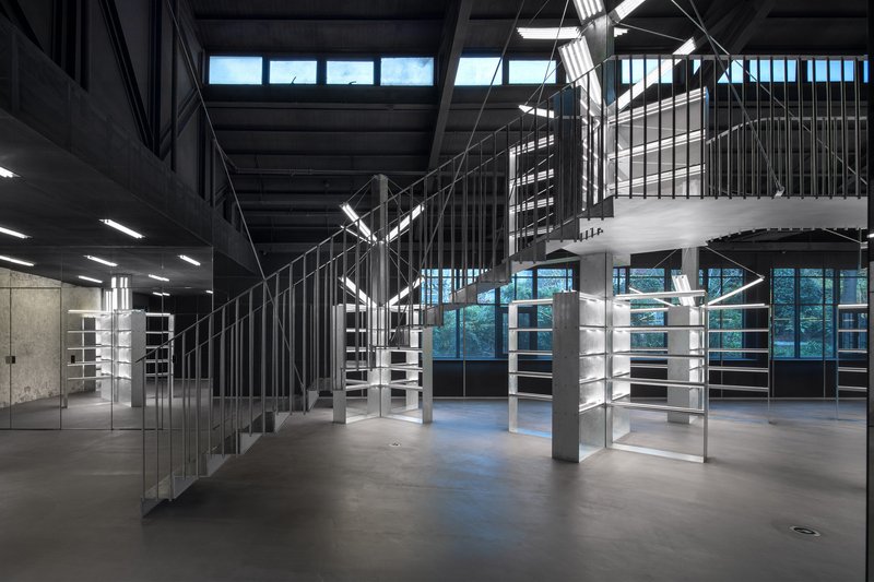 Double height retail space with freestanding glass shelving units and suspended stairs visible at dusk