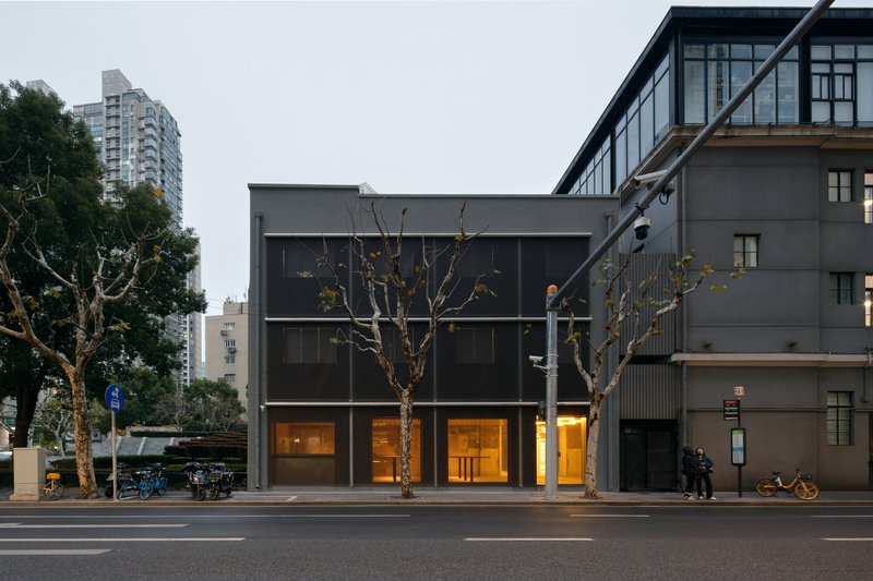 Street view of the grey box facade with pedestrians gathered near bare trees at twilight