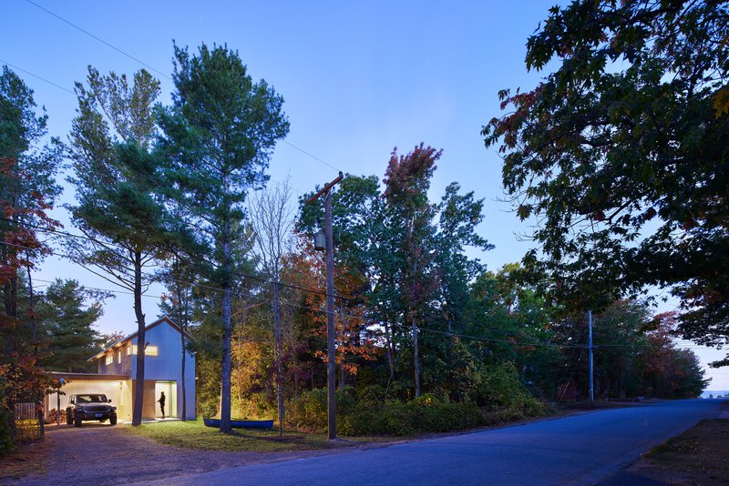 Street view of the gabled volume nestled among trees with illuminated windows at twilight